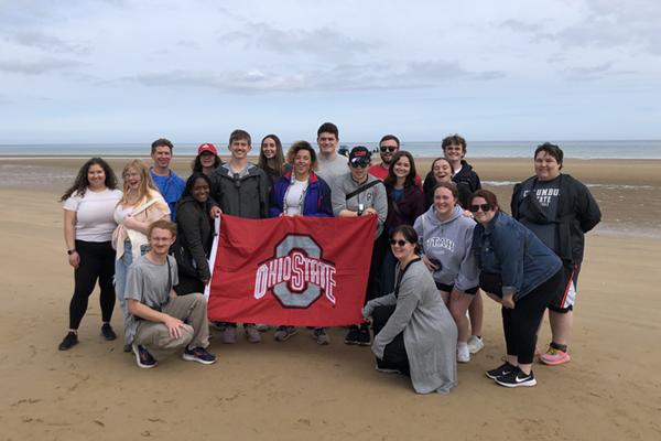 2024 WWII Study Tour Students standing on Omaha Beach holding an Ohio State flag